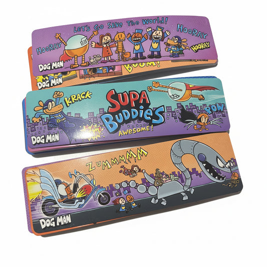 Three colorful metal dog toy paddles with cartoon graphics and text, stacked vertically on a light surface. The paddles feature characters and words like 'Dog Man', 'Krack', 'Supa Buddies', and 'Zummma'.